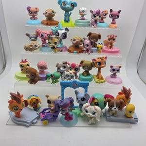May include: A collection of numerous small, colorful plastic animal figurines with large eyes, arranged on a four-tiered clear display stand. The figures include various species such as dogs, cats, birds, and mythical creatures in diverse poses.