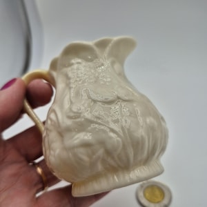 Peut inclure: Un petit pot à crème en céramique de couleur crème, orné d'un motif floral en relief sur tout son corps arrondi. Le pichet présente une anse incurvée, un bord festonné et une base évasée, avec une finition brillante qui reflète la lumière.