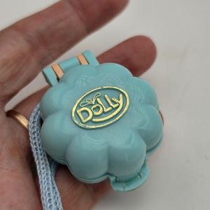 Peut inclure: Un boîtier compact en plastique bleu clair en forme de fleur avec un couvercle à charnière. Le centre présente un emblème ovale doré avec le mot DOLLY imprimé dans une police stylisée. Un cordon tissé bleu clair est fixé sur le côté.