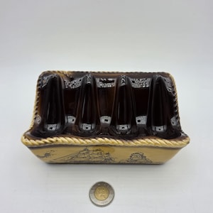 May include: A rectangular ceramic toast rack with a dark brown glazed top featuring four slots and a cream-coloured base decorated with a black line drawing of a sailing ship and a person. A Canadian one-dollar coin is placed in front for scale.