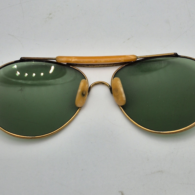 May include: Vintage aviator sunglasses with gold-tone frames and green lenses. The bridge features a tan bar, and the nose pads are a matching tan color. The lenses are oval-shaped, and the frames show signs of wear.
