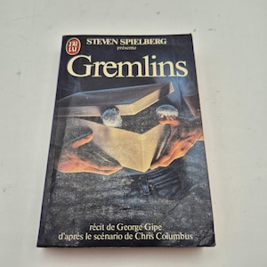 Peut inclure: Un livre de poche vintage intitulé "Gremlins" de Steven Spielberg. La couverture présente un fond bleu foncé avec le titre en grandes lettres blanches. L'illustration de la couverture représente une boîte avec deux petites créatures à l'intérieur.