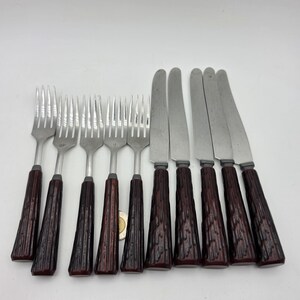 Könnte beinhalten: Ein Set aus fünf silberfarbenen Metallgabeln und vier silberfarbenen Metallmessern mit dunkelbraunen, strukturierten Griffen, die an Baumrinde erinnern. Das Besteck ist in einer Reihe angeordnet und zeigt die Metallzinken und Klingen.