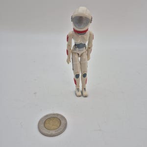 Könnte beinhalten: Eine kleine, cremefarbene Astronauten-Actionfigur mit weißem Helm, roten Akzenten und einem grauen Visier. Die Figur hat bewegliche Gelenke und trägt einen Raumanzug mit blauen und roten Details. Eine Münze befindet sich im Vordergrund.