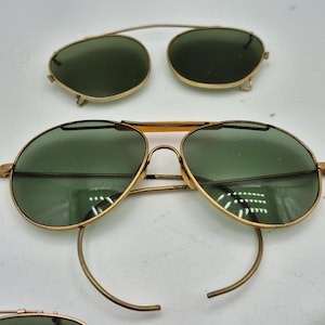May include: Vintage gold-tone aviator sunglasses with green lenses. Includes a separate clip-on pair. The sunglasses have a classic teardrop shape and a thin metal frame. The lenses are a dark green color.