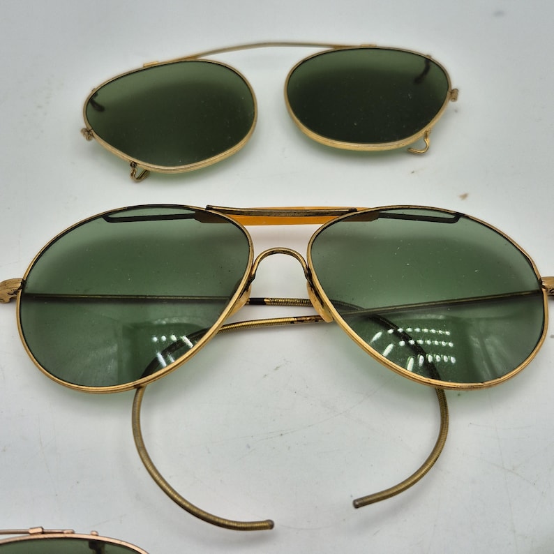May include: Vintage gold-tone aviator sunglasses with green lenses. Includes a separate clip-on pair. The sunglasses have a classic teardrop shape and a thin metal frame. The lenses are a dark green color.
