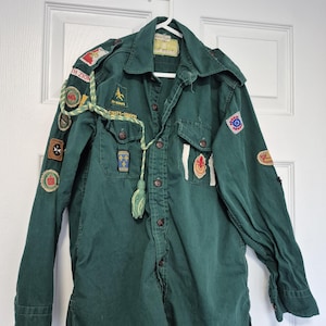Peut inclure: Une chemise de scout verte foncée avec plusieurs écussons et une corde décorative. La chemise a une fermeture boutonnée sur le devant, deux poches de poitrine et des manches longues. Les écussons comprennent du texte et divers emblèmes.