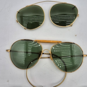 May include: Vintage gold-tone sunglasses with green lenses. The image shows two pairs of sunglasses: one complete pair and a clip-on pair. The lenses are round and the frames are thin. The clip-on pair has a bridge that attaches to the top of the glasses.