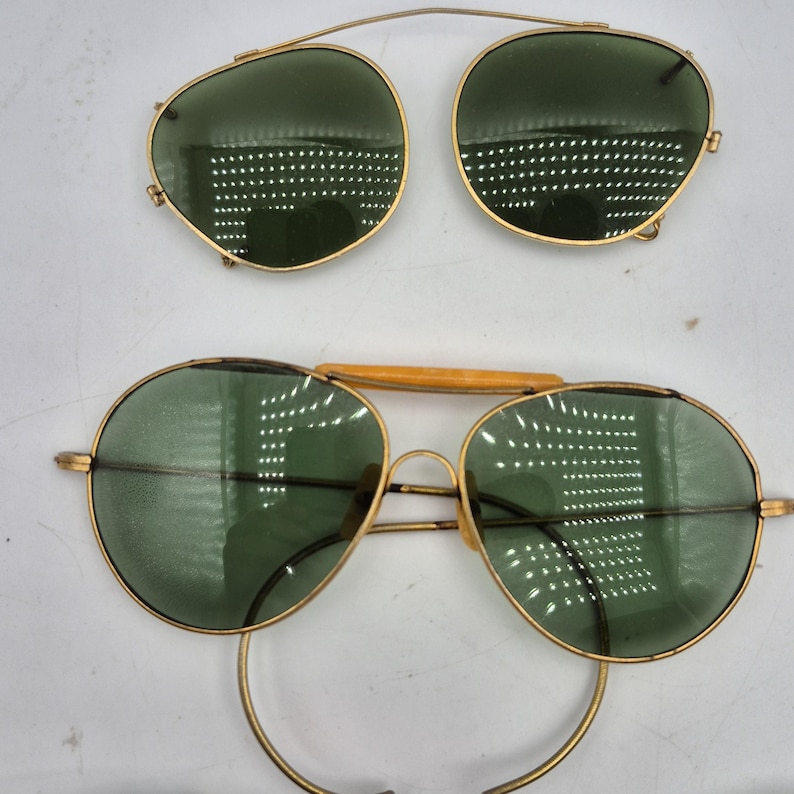May include: Vintage gold-tone sunglasses with green lenses. The image shows two pairs of sunglasses: one complete pair and a clip-on pair. The lenses are round and the frames are thin. The clip-on pair has a bridge that attaches to the top of the glasses.