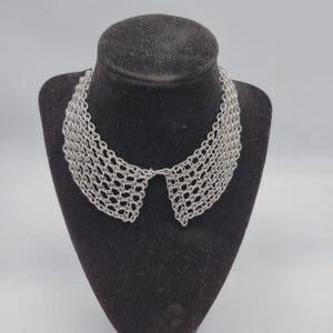 May include: Silver metal collar necklace with a woven, openwork design. The necklace is displayed on a black mannequin bust. The collar shape is reminiscent of a Peter Pan collar.