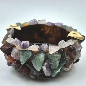 May include: A circular bowl featuring an exterior surface covered in a mosaic of tumbled gemstones including green aventurine, purple amethyst, clear quartz, and dark brown stones, accented with small gold-toned metal pieces along the rim.