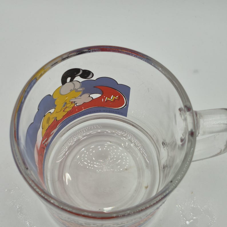 May include: Clear glass mug with a handle, featuring a cartoon graphic of a yellow character with a black head and a red lightning bolt. The graphic is set against a blue and white background.