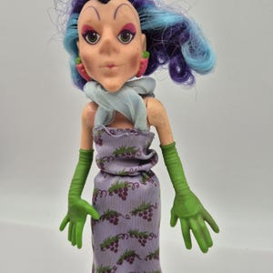 Peut inclure: Une poupée aux cheveux violets et bleus, portant une écharpe bleu clair, une robe lavande à motif de raisins violets et de longs gants verts. La poupée a des boucles d'oreilles roses en forme de carotte, des traits peints et un menton pointu.
