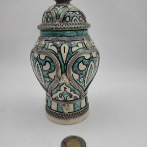 Peut inclure: Un pot décoratif en céramique avec un couvercle, présentant un motif turquoise et blanc avec des accents métalliques argentés. Le pot a une forme bulbeuse et un design détaillé. Le pot mesure environ 13 cm de haut, mélangeant couleurs et textures.