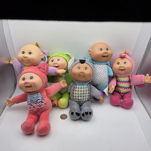 Peut inclure: Un ensemble de cinq poupées Cabbage Patch Kids. L'une porte une capuche de chat rose avec un torse à motif zébré, une autre une capuche verte, une capuche d'animal grise à pois, une tenue bleue à pois et une capuche rose à motif chevron.