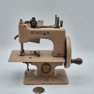 Puede incluir: Máquina de coser Singer vintage de color beige. La máquina tiene una manivela, una pequeña superficie de trabajo y el logotipo de Singer. Se encuentra sobre una base beige con un emblema circular. Una moneda está en primer plano.