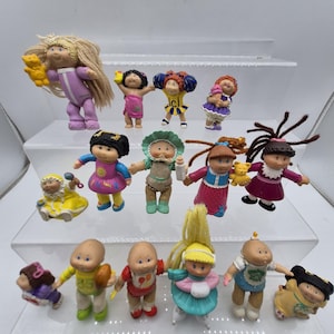May include: A collection of vintage Cabbage Patch Kids dolls, each with unique outfits and hairstyles. The dolls are in various poses, some holding accessories like teddy bears and bottles. The dolls are displayed on clear, tiered shelves.