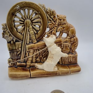 Könnte beinhalten: Eine braune Keramikfigur mit einem Hund an der Leine, einem Spinnrad und einer Katze. Der Hund ist cremefarben, und die Katze sitzt auf einer Mauer. Das Spinnrad ist ein wichtiges Merkmal. Die Basis ist rechteckig.