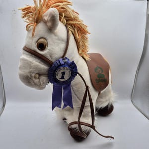 May include: A soft toy horse in white with a brown saddle and bridle. The horse features an orange and yellow mane and a blue ribbon with the text "Premier de la Parade" and the number 1.