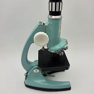 Peut inclure: Un microscope en métal bleu clair doté d'une platine noire, d'une molette de mise au point blanche et d'un oculaire marqué 10X. L'instrument comprend une tourelle porte-objectifs argentée et une base robuste pour l'observation scientifique.