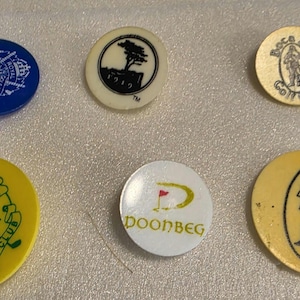 May include: Six circular golf ball markers featuring various designs and logos. The collection includes a blue marker, a cream marker with a tree, a tan marker labeled Boca Rio Golf Club, a yellow Elm Ridge Country Club marker, a white Doonbeg marker, and a tan marker with a figure.