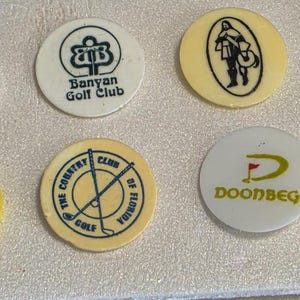 May include: A collection of twelve circular golf ball markers in various colors including white, yellow, and blue. Each marker features unique printed logos, emblems, and text such as Banyan Golf Club, Doonbeg, and Old Head, representing different golf courses.