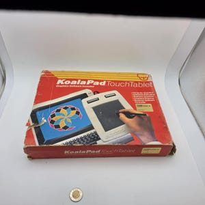 Puede incluir: Una caja vintage de KoalaPad TouchTablet con gráficos en rojo y blanco, texto que incluye 'KoalaPad TouchTablet' y 'Graphics Software Included', y una ilustración de una pantalla de ordenador y tableta. Una pequeña moneda de plata está delante.