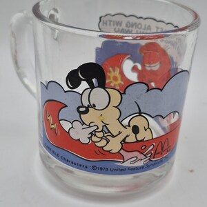 May include: Clear glass mug featuring Garfield characters. The mug has a cartoon illustration of Garfield in a boat with a red wave and a sun. The mug has a handle and text that reads "Garfield Characters: &copy;1978 United Feature Syndicate".