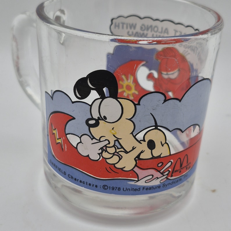 May include: Clear glass mug featuring Garfield characters. The mug has a cartoon illustration of Garfield in a boat with a red wave and a sun. The mug has a handle and text that reads "Garfield Characters: &copy;1978 United Feature Syndicate".