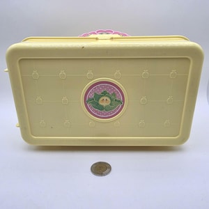 Peut inclure: Une mallette de transport rectangulaire en plastique jaune pâle ornée d'un motif en grille de petites pommes en relief. Un emblème circulaire rose et blanc à pois au centre affiche un personnage de chou. Une pièce d'argent est placée devant.