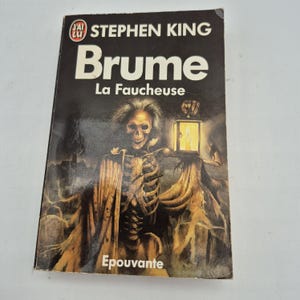 Peut inclure: Un livre de poche intitulé "Brume" de Stephen King. La couverture présente un squelette tenant une lanterne, avec les mots "La Faucheuse" et "Epouvante". L'arrière-plan est sombre avec un thème effrayant.