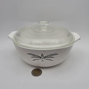 May include: A white round casserole dish with integrated side handles and a clear glass lid. The exterior features a black wheat stalk graphic. A small circular coin is placed in front of the dish for scale, showing the relative size of the item.