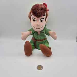 May include: A Peter Pan plush doll, dressed in a green tunic, brown shoes, and a red feather in his brown hair. The doll has a smiling face and outstretched arms. A coin is in the foreground.