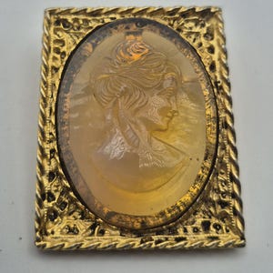 Peut inclure: Une broche rectangulaire dorée avec un camée ovale central en résine de couleur ambre. Le camée représente le profil d'une personne avec une coiffure relevée complexe. Le cadre présente une bordure texturée en corde et un motif ajouré orné.