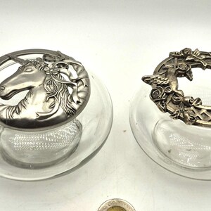 May include: Two clear glass jars with decorative silver-toned lids. One lid features a unicorn head, the other an oval design with roses. The jars are on a white surface.