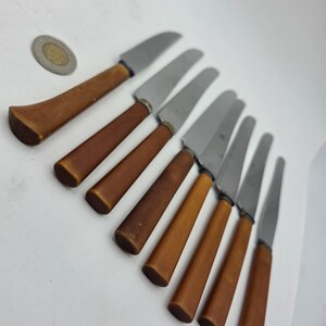 May include: A set of seven knives with silver-toned metal blades and smooth, light brown handles arranged in a diagonal row, accompanied by a single circular coin placed near the top left corner for scale reference.
