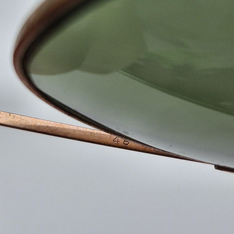 May include: Close-up of sunglasses with a green lens and a gold-colored frame. The number "48" is printed on the frame. The sunglasses have a classic design.