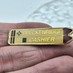May include: A rectangular gold-tone metal name badge pin featuring a cream-colored background with the text BRECKENRIDGE and CASHIER in gold lettering. The left side displays a small black square and a decorative four-petal floral symbol.