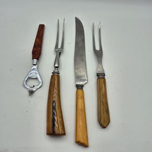Può includere: Una collezione di utensili da portata vintage, tra cui un apribottiglie, due forchette da intaglio e un coltello da intaglio. Gli utensili presentano manici in legno con diverse tonalità di marrone e beige e lame e teste in acciaio inossidabile.