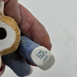 Peut inclure: Une figurine en céramique avec une tête texturée beige et un pantalon bleu. Le dessous du pied porte l'inscription 1984 OAA, INC. 64EDITION TAIWAN. La figurine présente une ouverture circulaire creuse sur le dessus de la tête et une base plate.