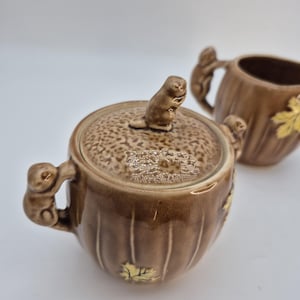 Puede incluir: Juego de azucarero y crema de cerámica marrón. El azucarero tiene una tapa texturizada con un asa en forma de ardilla. Ambas piezas presentan asas de ardilla y detalles de hojas. La crema está en el fondo.