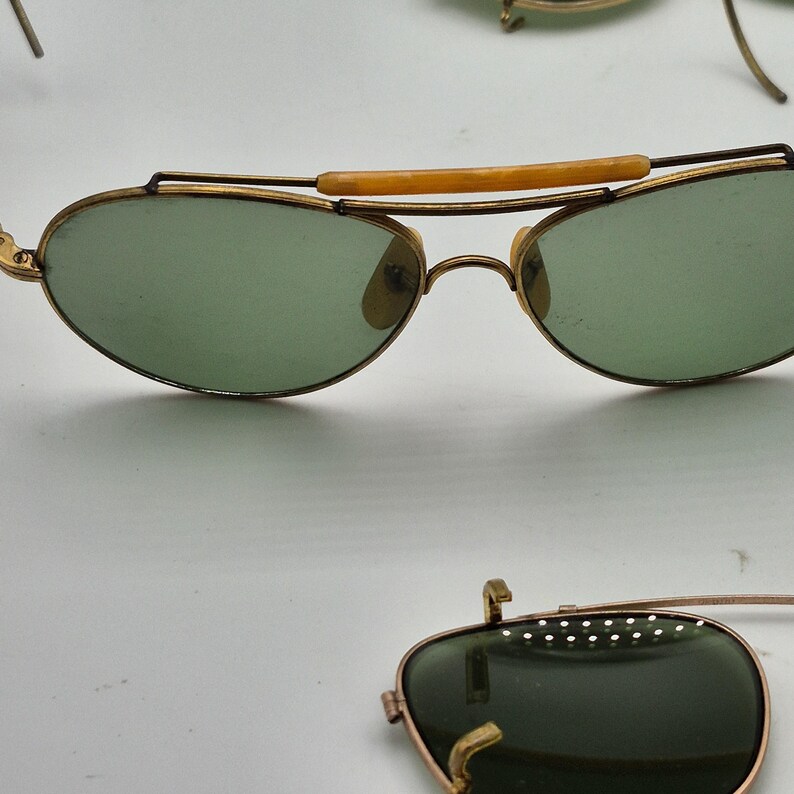 May include: Vintage aviator-style sunglasses with gold-tone frames and green lenses. The sunglasses feature a bridge bar and a light brown brow bar. The lenses are oval-shaped. The image shows multiple pairs of sunglasses.