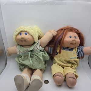 Pode incluir: Duas bonecas com corpos de tecido macio e cabeças de vinil. A boneca da esquerda tem cabelo de lã loiro, olhos azuis e um vestido xadrez verde. A boneca da direita tem cabelo ruivo com mechas roxas, olhos azuis e jardineiras de veludo amarelo.