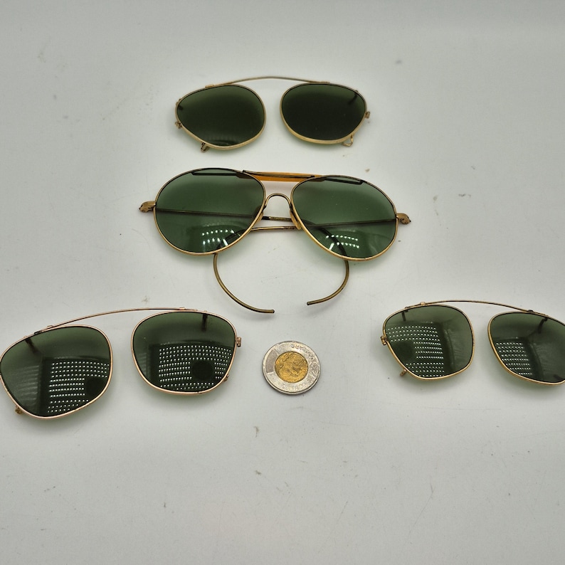 May include: An assortment of vintage sunglasses and clip-on lenses with gold-colored frames and green lenses. The sunglasses are in various styles, including aviator and clip-on designs. A Canadian coin is also present.