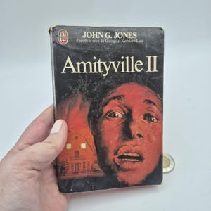 Peut inclure: Un livre de poche vintage intitulé "Amityville II" de John G. Jones. La couverture présente un portrait déformé et teinté de rouge d'une personne avec une expression terrifiée et une maison avec des fenêtres éclairées. Le livre est tenu par une main.