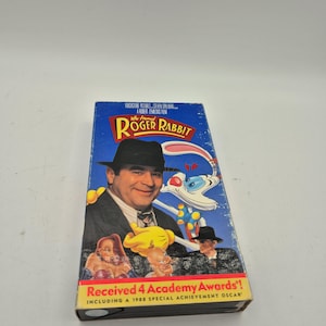 Peut inclure: Une cassette VHS du film "Qui veut la peau de Roger Rabbit" avec un fond bleu. La couverture présente le titre du film, des personnages et le texte "Received 4 Academy Awards!"