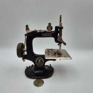 Peut inclure: Une machine à coudre miniature Singer vintage, noire avec des détails argentés. La machine comprend un volant, une aiguille et une petite base. Le mot "SINGER" est visible sur le bras. Une pièce de monnaie est placée devant la machine.