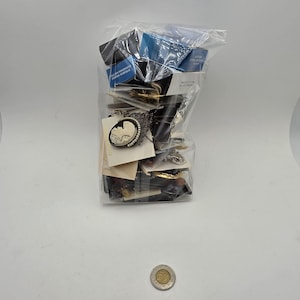 Peut inclure: Un sac en plastique transparent rempli d'un assortiment de bijoux, incluant une broche camée noire et blanche avec une bordure de perles, diverses boîtes et cartes. Une pièce de monnaie argentée est placée devant le sac pour servir d'échelle.