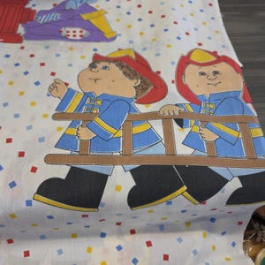 May include: White fabric featuring a repeating pattern of cartoon characters dressed as firefighters in blue uniforms with red helmets, carrying a brown ladder. The background is decorated with small, scattered red, blue, and yellow squares.
