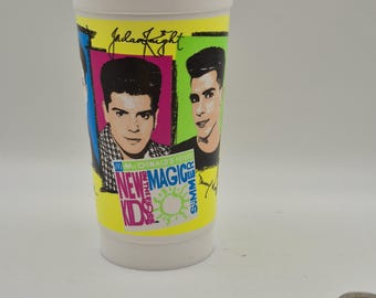 Puede incluir: Un vaso de plástico blanco con una banda amarilla que presenta imágenes de dos hombres y el texto "New Kids on the Block". La taza también tiene el texto "McDonald's Show" y "Summer".